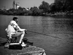 Mann beim Angeln am Flussufer in Sevilla, sitzend auf einem Campingstuhl mit Angelrute, Schwarzweiß-Fotografie mit Industriegebäuden im Hintergrund
