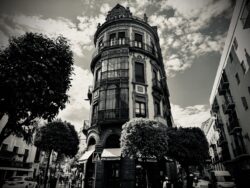 Rundes Jugendstilgebäude in Sevilla von unten fotografiert mit Bäumen im Vordergrund und bewölktem Himmel, Schwarzweiß-Architekturstreetstyle