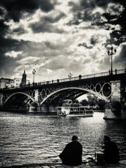 Puente de Triana über den Guadalquivir in Sevilla mit dramatischem Wolkenhimmel, zwei Silhouetten im Vordergrund, Schwarzweiß-Fotografie