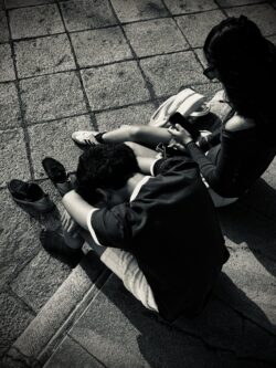Zwei junge Personen von oben fotografiert, sitzend auf Pflastersteinen, beide mit Smartphones, hartes Seitenlicht, Schwarzweiß-Streetfoto Sevilla