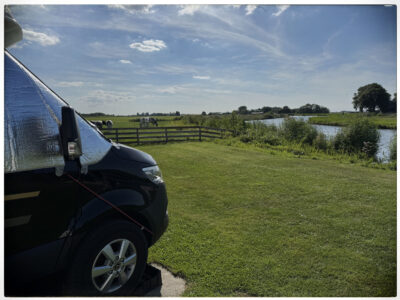 Blick von der Motorhaube des Wohnmobils auf eine grüne Wiese mit weidenden Kühen und einen Flusslauf in Friesland.