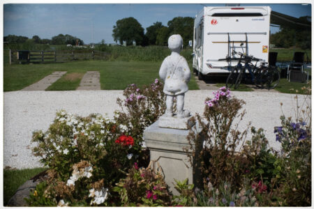 Eine kleine weiße Steinfigur eines Jungen steht in einem bunten Blumenbeet auf dem Stellplatz, im Hintergrund ein Wohnmobil.
