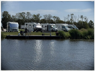 Aufnahme vom Wasser aus auf eine Reihe geparkter Wohnmobile am Ufer des Camperplaats aan de Ee bei Dokkum.