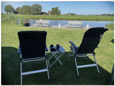 Zwei Campingstühle stehen auf der Wiese mit Blick auf das Wasser, auf dem Beistelltisch stehen Kaffeetassen und eine Kamera.