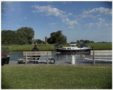 Eine Person sitzt entspannt auf einer Holzbank an einem Anlegesteg, während ein Motorboot auf dem Kanal vorbeifährt.