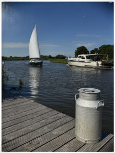 Eine alte silberne Milchkanne steht auf einem Holzsteg am Kanal, auf dem Wasser fahren ein Segelboot und ein Motorboot.