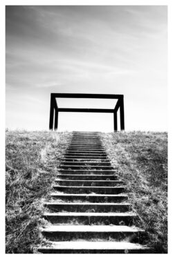 Treppe führt zu einem freistehenden schwarzen Stahlrahmen auf der Deichkuppe, minimalistische Komposition vor hellem Himmel, Schwarzweiß
