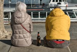 Rückenansicht eines Paares, das mit einer Flasche Bier am Kölner Rheinufer sitzt.