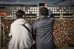 Zwei Männer sitzen an der Hohenzollernbrücke und fotografieren einen vorbeifahrenden Zug mit dem Smartphone.