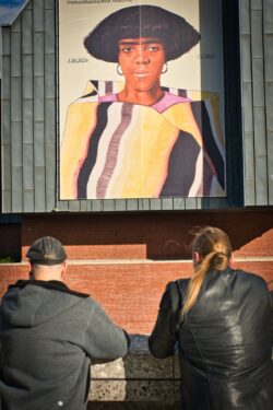 Zwei Menschen von hinten betrachten ein großes Kunstplakat an einer Fassade.